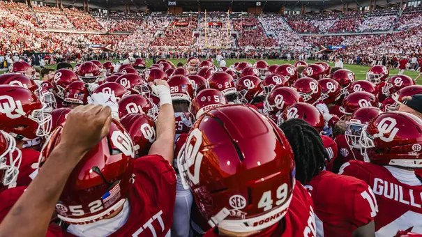 Oklahoma football team huddle photo showing red helmets and jerseys with arms raised and crowd visible in background