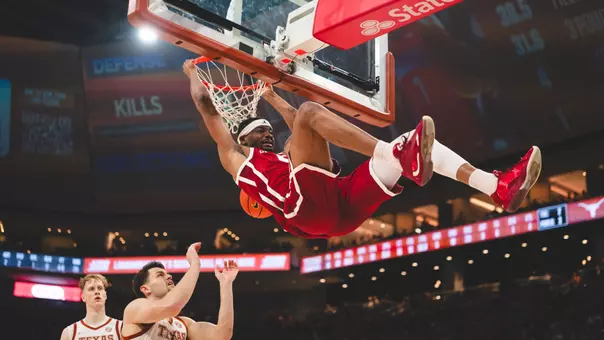Mohamed Wague in red uniform kicks his legs up at the end of an emphatic dunk in front of two Texas defenders in white uniform in OU's win at Texas at Moody Center in Austin, Texas, on March 7, 2026.