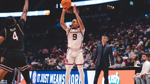 Nijel Pack in white uniform shoots a 3-pointer over a South Carolina defender, in black uniform, as OU head coach Porter Moser looks on in the background, during OU's SEC Tournament win during the SEC Tournament at Bridgestone Arena in Nashville, Tenn.