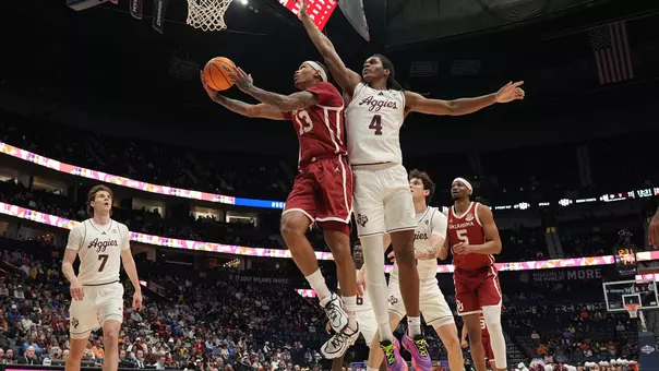 OU's Tae Davis in red uniform goes up for a layup against a Texas A&M defender in white uniform as players from both teams look on, in the SEC Tournament at Bridgestone Arena in Nashville, Tenn.