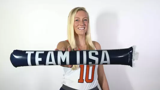 Charlotte de Vries stands on a white backdrop with a white Syracuse field hockey jersey on. It has her #10 in a big Orange font on the front. In her extended right arm is a blue thunderstick noise maker with TEAM USA in white font.