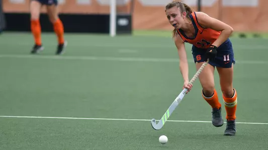 Claire Cooke dribbles the ball up field. She has a concentrated look on her face while wearing an orange sleeveless jersey and a dark blue skirt. Her socks are Syracuse orange, with a yellow and white Captains band around her left shin guard.