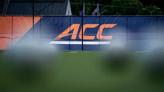 IN big orange letters is the ACC logo on a windscreen. The logo is on a blue background with an orange portion to the left on an angle. Sitting on a green turf field are several field hockey balls, artfully out of focus to display depth of field.