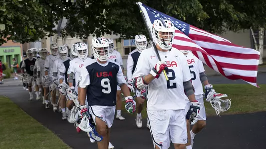 Team USA Lacrosse member Cole Kirst leads the team onto the championship field in Limerick. He is carrying an American flag behind him, gracefully, laid across his shoulders from the breeze. His compatriots walk in two lines behind him, all sport white Team USA kit with white helmets, featuring a blue stripe with white stars and an alternating red and white stripe on the other side.