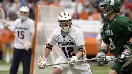 Syracuse defenseman Kyle Guadagnolo eyes his opponent from Loyola. Guadagnolo is wearing an all-white Syracuse university with SYRACUSE and his number 42, on the front in dark blue. His opponent wears an all kelly green kit. A freshman John Galloway stands in the background guarding his net.