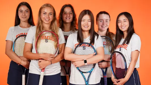 All six members of the 2022-23 Syracuse tennis team stand together on an Orange background. They're wearing white Syracuse shirts, holding their tennis rackets in front of them.