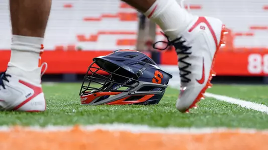 A Syracuse University men's lacrosse helmet rests on the turf of the JMA Dome. A player walks by with a pair of white Nike cleats.