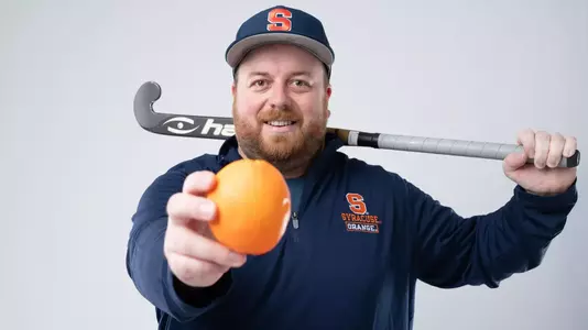 Syracuse field hockey assistant coach Sam Brown stands with a field hockey stick across his shoulder, holding an Orange towards the camera. He's wearing a blue pullover with Syracuse University branding and a blue hat all on a white background.