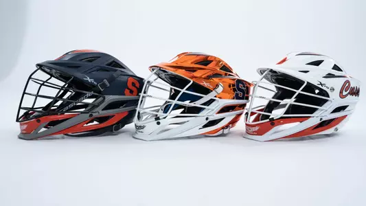 Three Cascade lacrosse helmets lined up on a white background. Matte Blue, Chrome Orange, and white shells from left to right.
