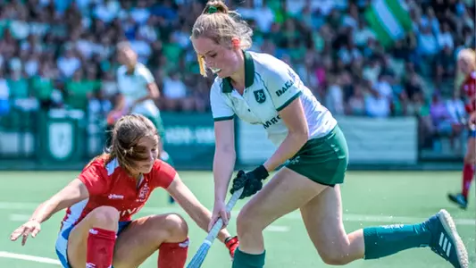 Cato Schreinemacher, wearing a white and green field hockey kit, dribbles past a would-be defender in red, who is falling down in awe of Schreinemacher's talent.