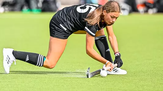 Bo Madden, wearing an all-black field hockey kit, sweeps her stick along the water-soaked turf field, spraying up water before striking the ball.