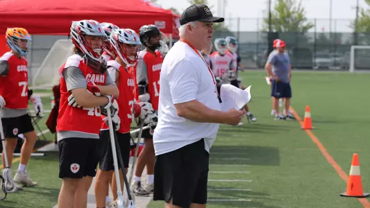 Gary Gait patrols the sidelines of a Canada Lacrosse U20 development camp. Gait wears a white shirt, black shorts and black baseball cap. Canada Lacrosse players stand behind him.