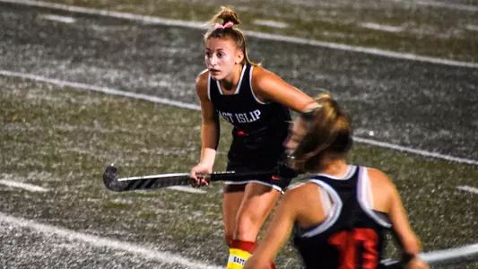 Sarah Smalley on the field hockey field during a night game. She wears a black East Islip High School uniform with a gold and white captains band around her red shin guards.