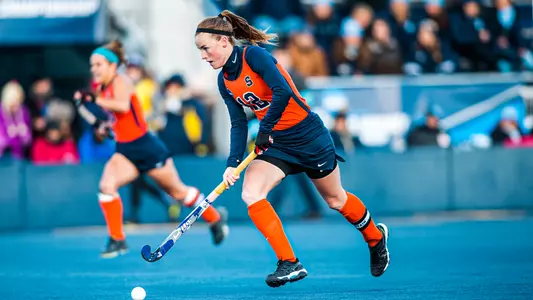 Emma Russell dribbles down field during the DI national championship game in 2015. The astro turf is blue at Michigan's field, while Russell wears an Orange tank and blue kilt, with a blue longsleeve shirt on under the tank.