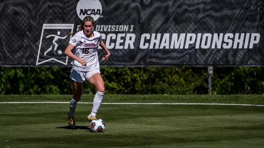 Macie Kolb, in the Aggies' all-white kits, is on the dribble attack with a monstrous 2020 NCAA Division I Soccer Championship banner in the back ground at Macpherson Stadium in Greensboro, North Carolina