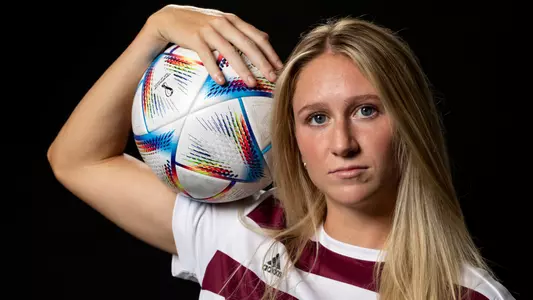 Kate Colvin in front of a black background on portrait day, flexing her right arm with a soccer ball in tow.