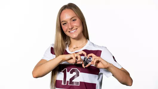 Ali Russell making a hand heart above the Texas A&M logo on her white with maroon stripes soccer jersey in front of a white background.