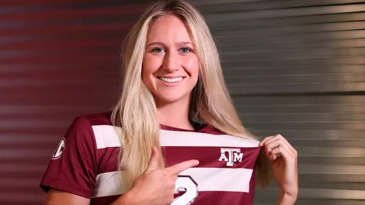 Kate Colvin in a portrait shoot in front of a metal corrugated wall pointing at the Texas A&M crest on her maroon jersey
