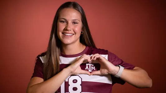 Macy Matula drops heart hands over the Texas A&M logo on her maroon jersey in front of a maroon portrait background.
