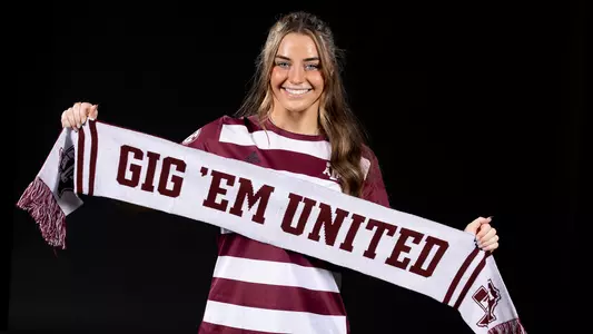 Shae O'Rourke poses at her recruiting trip portrait shoot holding a smartly elegent gray Gig'Em United scarf while wearing an Aggie maroon uniform in front of a stark black background.