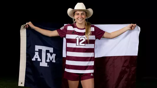 Leah Pirro poses with a cowboy hat and a Lone Star style Texas A&M flag while wearing an Aggie maroon jersey in front of a stark black background.