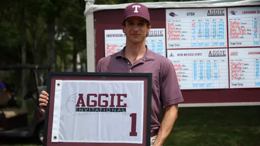Jaime Montojo dressed in a class maroon polo with vertical white pinstripes while donning the popular maroon hat with a white block T posses with the Aggie Invitational prize, a framed pin flag.