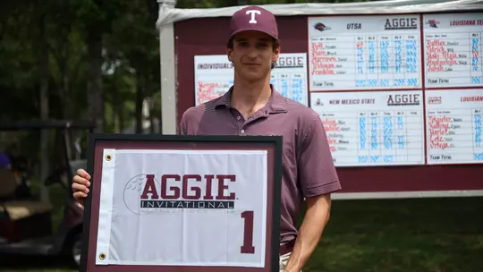 Jaime Montojo dressed in a class maroon polo with vertical white pinstripes while donning the popular maroon hat with a white block T posses with the Aggie Invitational prize, a framed pin flag.