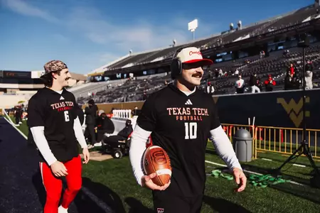 Texas Tech player in black warm-up gear walks along the sideline holding a football while another player follows behind.