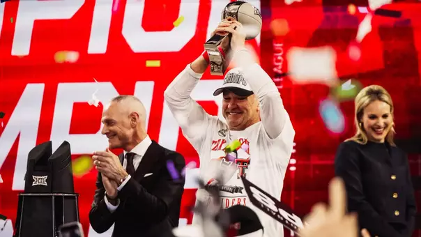 Texas Tech head coach Joey McGuire smiles and raises the Big 12 championship trophy overhead on the podium as Big 12 Commissioner Brett Yormark applauds beside him with confetti falling in front of a red championship backdrop.