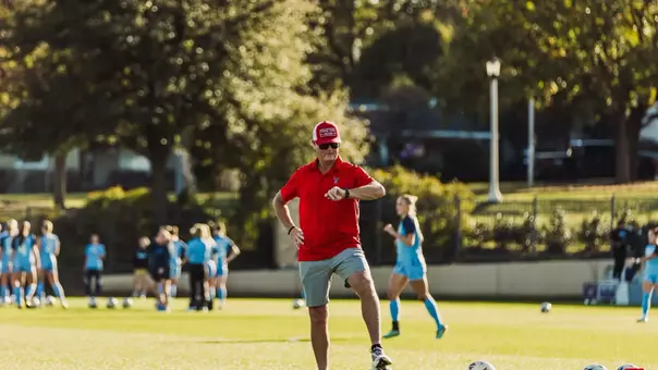 Tom Stone stares at his wrist watch, while leaning on a Wilson Soccer ball and dressed sharply in a Red Texas Tech Soccer hat, Red Double-T adorned polo and gray golf shorts prior to the Red Raiders NCAA Round of 32 match against the University of North Carolina at TCU back on Nov. 19, 2025 on a picture-perfect late autumn afternoon in Forth Worth