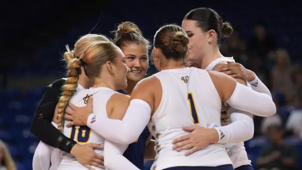 Members of the UC Irvine women's volleyball team wearing white jerseys and blue shorts huddle up on the court.