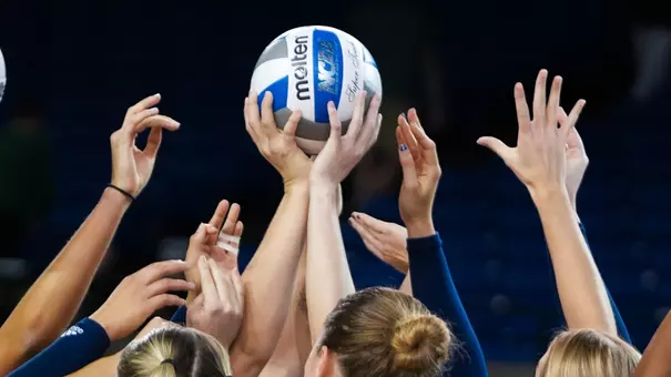 Women's volleyball players huddle while holding a ball up in the middle