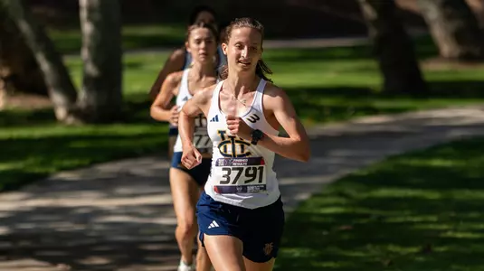 Anna Soares wearing a white UCI jersey and blue shorts running a cross country course at a park.