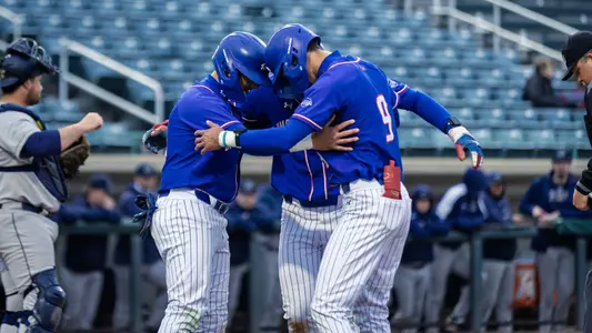 UMass Lowell baseball players in blue uniforms celebrating at home plate after scoring a run in the contest