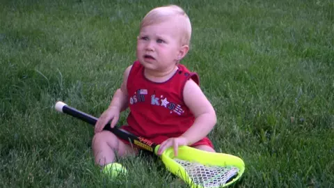 Lauren LaPointe as baby sitting in the grass holding a lacrosse stick