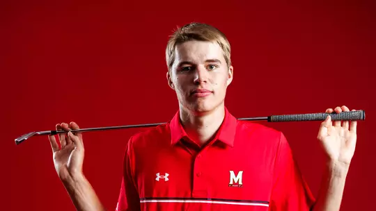 Studio photo of Emil Riegger with a golf club across his shoulders on a red background