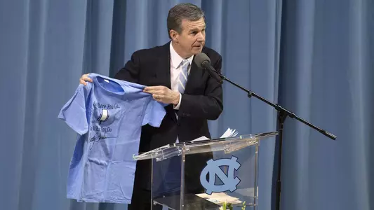 Governor Roy Cooper holds up a t-shirt with a Woody Durham quote while making remarks during the Woody Durham memorial service in Carmichael Arena, Sunday, April 8, 2018 in Chapel Hill, N.C. The former play-by-play announcer died at the age of 76 on March 7. (UNC Athletic Communications/Jeffrey A. Camarati).