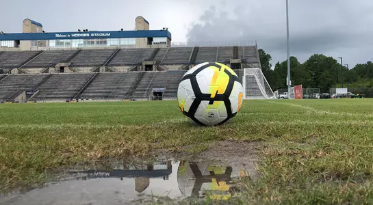 A soccer ball rests next to the corner flag and a puddle of water with Hodges Stadium in the background