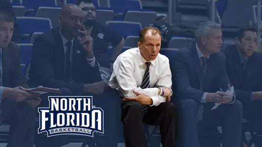 Coaching staff sits on the bench with a UNF basketball logo in the lower left corner