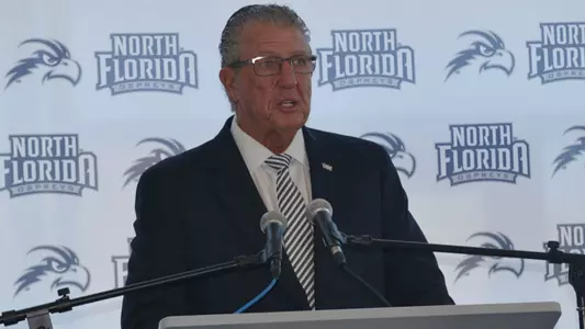 Lee Moon stands behind podium in front of a UNF Athletics backdrop