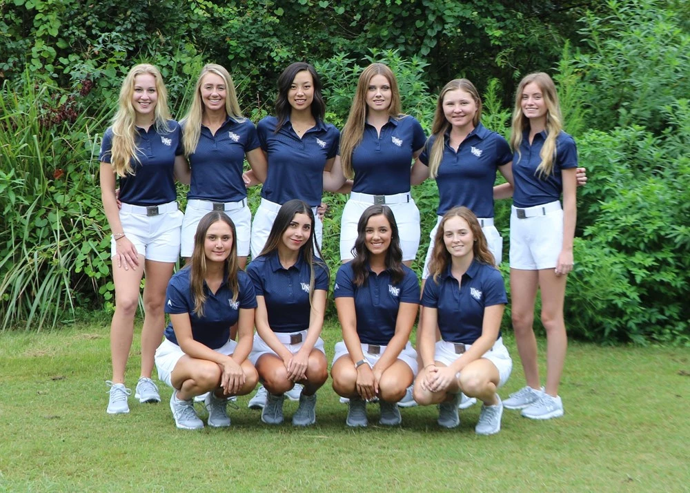 Women's golf team poses in two lines in front of a wall of bushes