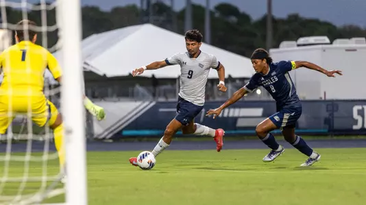 Forward Geraldo Neto dribbles a ball into the goal area with an opposing goalkeeper and player in pursuit