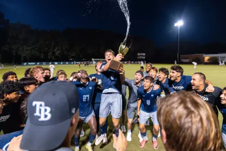 UNF Men's soccer team celebrates a win by lifting up a trophy together.