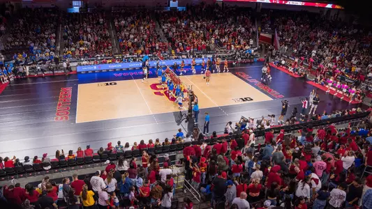 A high stadium shot of a large crowd at Galen Center during the 2024 USC-UCLA women's volleyball match. The teams are lined up to shake hands at center court on either side of the net.