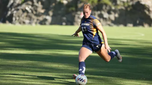 A soccer player in blue on a sunny day is kicking the ball during a game.