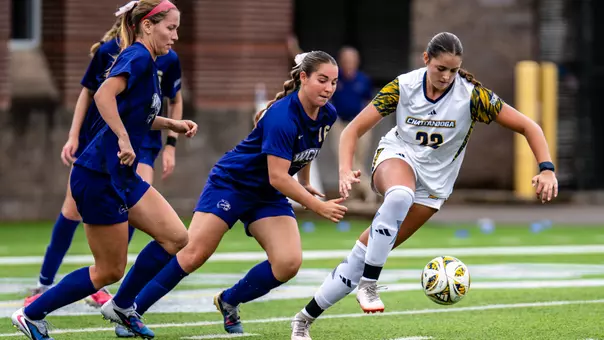 A Chattanooga soccer player wearing a white No. 22 jersey dribbles the ball on a turf field while two opposing players in blue uniforms close in from the left.