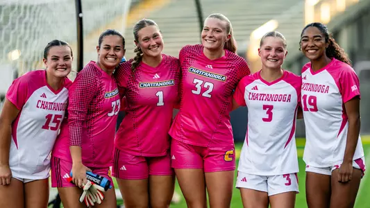 Six soccer players wearing a combination of pink kits and white kits with pink trim.