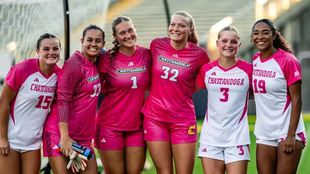 Six soccer players wearing a combination of pink kits and white kits with pink trim.
