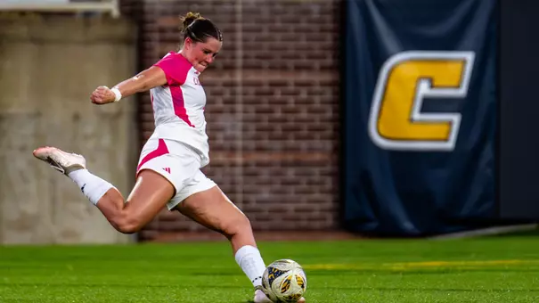 a soccer player in a white kit with pink trim is kicking the ball with a brick wall in the background.