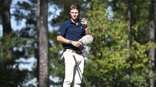 Evan Rogers musses his own hair with his hat in his hand at waist level awaiting his turn on the tee.